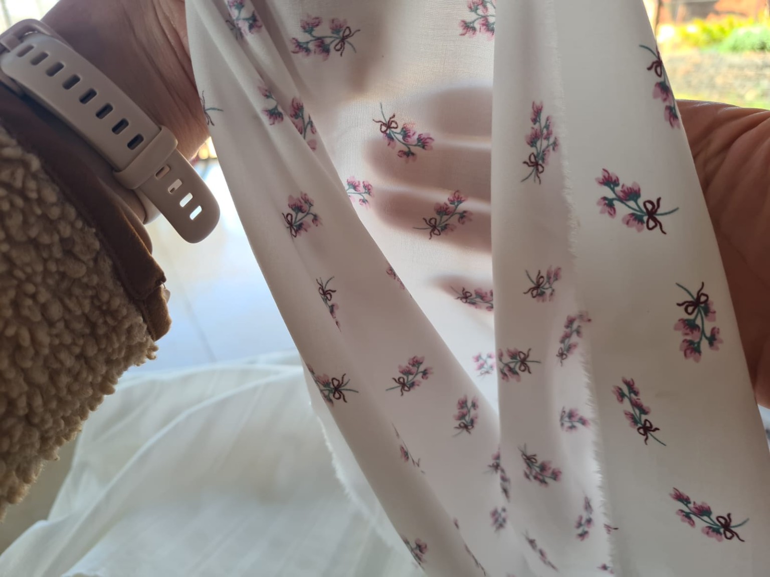Mãos segurando um tecido floral claro diante da luz, permitindo perceber a transparência e a mão por trás